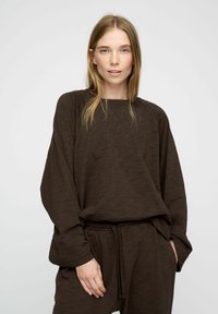 Barna, textúrált, oversize pulóver hosszú ujjal és kerek nyakkivágással, amelyhez laza szabású, húzózsinórral ellátott nadrág tartozik.