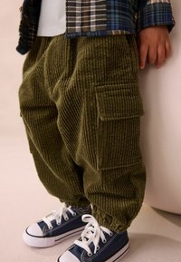 Olivgrüne Cordhosen mit gerippter Textur, ausgestattet mit seitlichen Taschen und elastischen Bündchen, kombiniert mit marineblauen Sneakers mit weißen Schnürsenkeln.