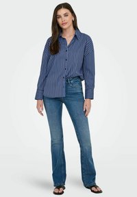 Chemise boutonnée rayée bleu et blanc avec un col, associée à un jean flare bleu clair et des sandales noires. Tissu lisse avec une coupe décontractée.