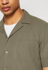 Camisa de manga corta en tejido texturizado color verde oliva, con cuello con muescas, un bolsillo en el pecho y cierre de dos botones.