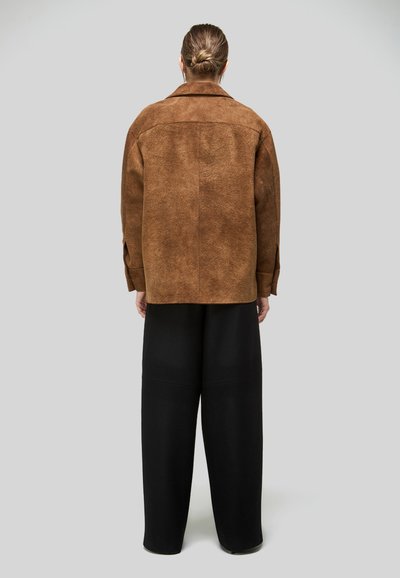 Kontyba fogott hajú személy barna szarvasbőr dzsekit és széles szárú fekete nadrágot visel, háttal áll egy egyszerű szürke háttér előtt.