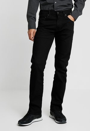 Mężczyzna ubrany w czarne slim-fit jeansy, czarne trampki i ciemnoszarą koszulę zapinaną na guziki, pozujący z jedną ręką w kieszeni.
