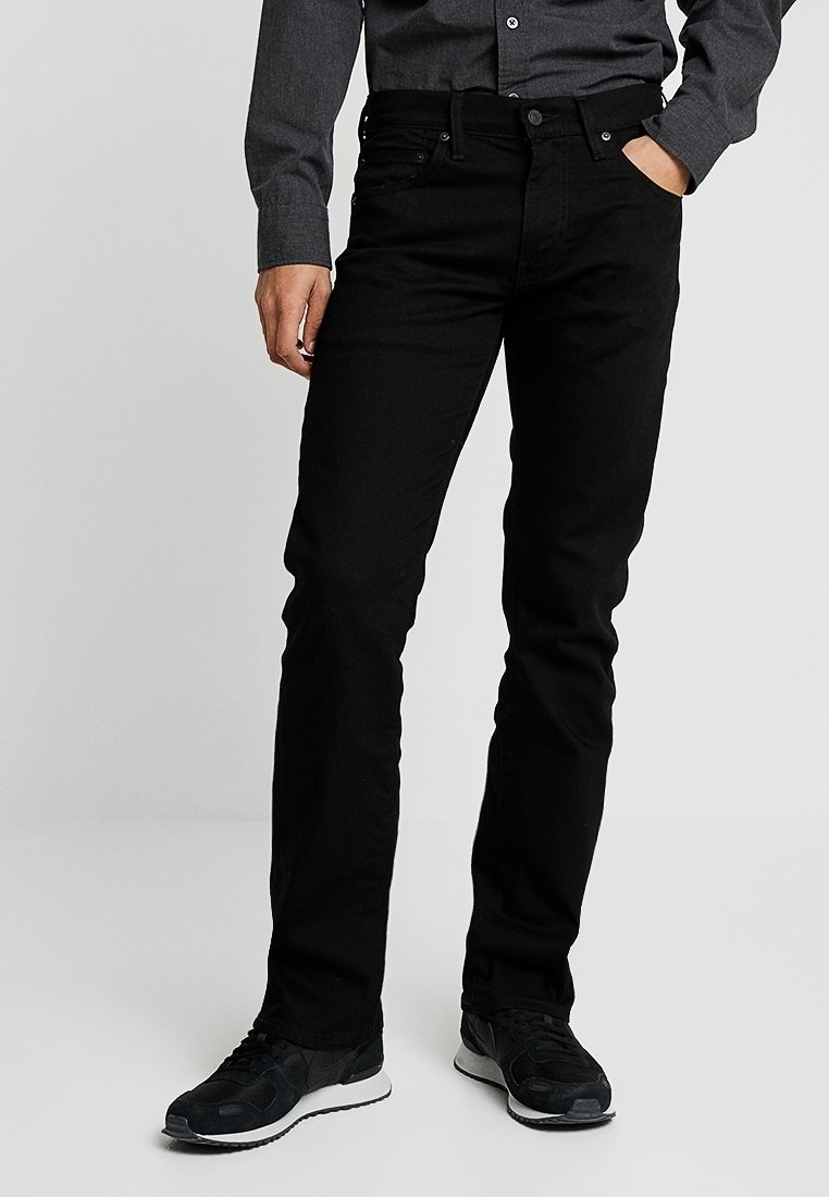 Homme portant un jean noir slim, des baskets noires et une chemise gris foncé boutonnée, posant avec une main dans la poche.