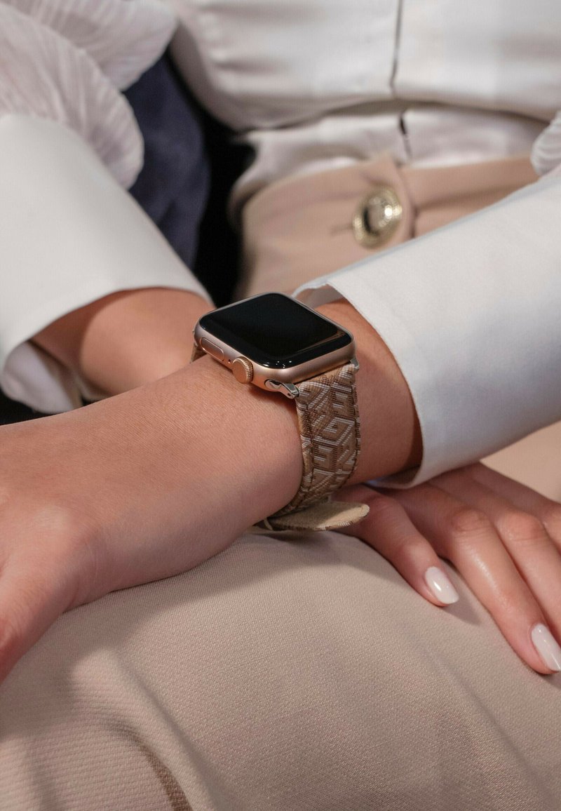 Smartwatch z kwadratowym czarnym wyświetlaczem i teksturowanym beżowym paskiem z tkaniny z geometrycznym wzorem, noszony na nadgarstku na jasnym tle.