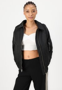 Veste bomber noire avec une texture lisse, fermeture éclair argentée et col, portée sur un haut court blanc ; le mannequin présente une tenue décontractée.