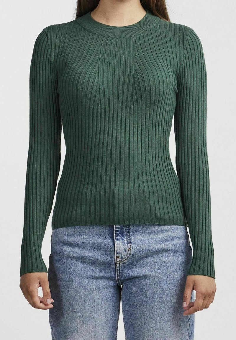 Femme portant un pull moulant à manches longues côtelé de couleur vert foncé, associé à un jean bleu clair, debout devant un fond uni.