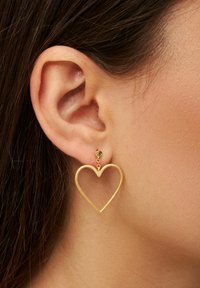 Orecchini a forma di cuore in oro appesi a uno stud, caratterizzati da una finitura testurizzata e una delicata catena. Posizionati sull'orecchio di un modello.