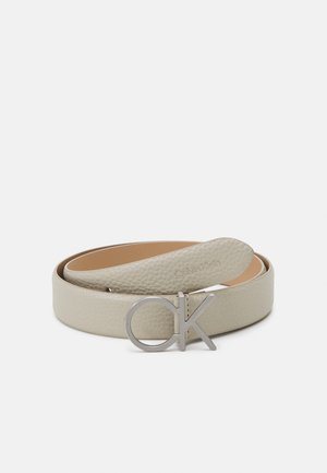 Jasnobeżowy, teksturowany skórzany pasek zwinięty na płasko z srebrną klamrą z logo „CK” Calvin Klein na białym tle.