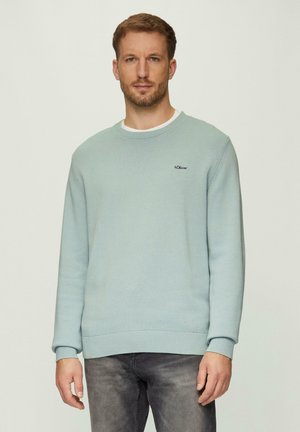 Helles blaues Strickpullover mit langen Ärmeln, gerippten Bündchen und Saum, mit einem kleinen Logo auf der Brust. Über einem weißen Rundhalshemd getragen.