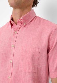 Camicia a maniche corte in tessuto a trama leggera di colore rosso chiaro, con un motivo testurizzato, colletto con bottoni e taschino sul petto. Presenta bottoni beige.