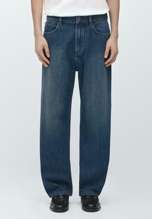 Personne portant un jean en denim bleu foncé à coupe ample avec un design classique à cinq poches et des chaussures en cuir noir, debout devant un fond uni.