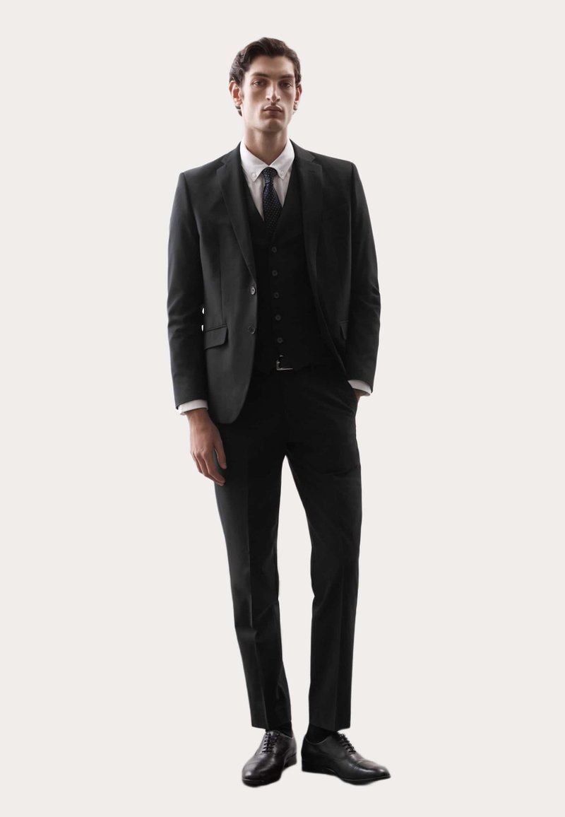 Schwarzer Anzug mit Slim-Fit, mit aufgesetztem Revers, weißem Hemd und einer Krawatte. Polierte schwarze Schuhe runden das Outfit ab.