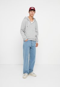Sudadera con cremallera en gris claro con capucha ajustable, combinada con vaqueros de pierna ancha en azul claro y zapatillas blancas. El fondo es completamente blanco.