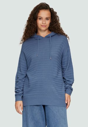 Un sweat à capuche bleu avec un design texturé et côtelé, équipé d'un cordon de serrage. Il présente une coupe décontractée, des manches longues et un ourlet large. Accompagné d'un pantalon en denim ample.