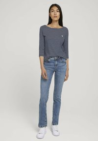 Haut à rayures marines et blanches à manches longues avec un col rond, associé à un jean bleu et des baskets blanches, se tenant devant un fond uni.