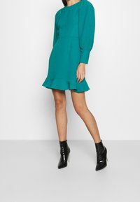 Robe turquoise avec manches longues bouffantes et ourlet évasé. Le modèle porte des bottines noires vernies. Texture lisse, silhouette structurée.