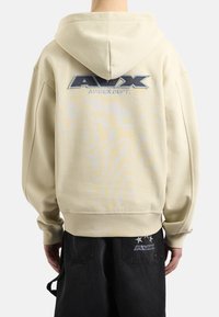 Felpa beige con cappuccio caratterizzata da un grande logo ricamato sul retro e polsini a costine. Indossata con pantaloni neri che presentano un logo più piccolo.