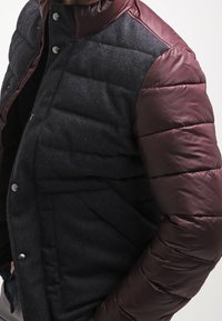 Båstad jacka med en svart strukturerad ullkropp och bourgogne släta syntetärmar. Inkluderar tryckknappar och stå-up krage.