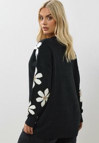 Schwarzer Strickpullover mit weißen Margeriten und beigen Zentren an den Ärmeln. Das Design umfasst gerippte Bündchen und unteren Saum, die eine lockere Passform bieten.