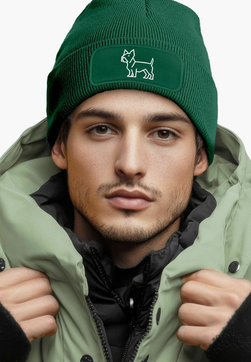 Green ribbed beanie with a dog design patch; worn with a light green puffer jacket; close-up of face and shoulders, neutral background.