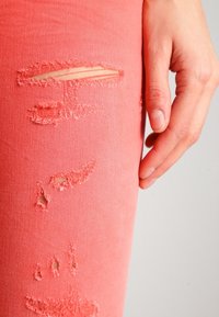 Mano descansando al lado de unos pantalones de mezclilla rasgados en color coral, que muestran múltiples desgastes en la tela.