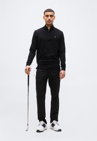 Άνδρας με μαύρο πουλόβερ με φερμουάρ στο λαιμό και παντελόνι κρατάει ένα γκολφ κλαμπ, στέκεται σε απλό λευκό φόντο, φοράει λευκά και μαύρα παπούτσια γκολφ.