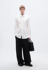 Λευκό πουκάμισο με κουμπιά και μακριά μανίκια, συνδυασμένο με φαρδιά μαύρη παντελόνια. Το μοντέλο κρατά μια μαύρη τσάντα και φορά μαύρα παπούτσια.