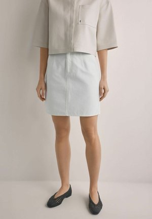 Kobieta w beżowym krótkim topie z kieszenią, białej spódnicy z wysokim stanem i czarnych plecionych balerinach stojąca na tle gładkiej ściany.