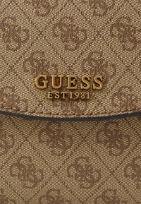 Nærbillede af en brun struktureret håndtaske med et gentagende monogrammønster og en guld "GUESS EST 1981" logo-plaque på frontklappen.
