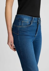 Jeans in denim blu con vita media, tasche frontali e una texture liscia, mostrati di lato, evidenziando la vestibilità e i dettagli delle cuciture.