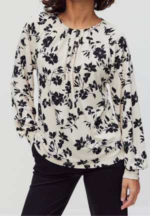 Kobieta ubrana w białą, długorękawową bluzkę z czarnym kwiatowym wzorem, wsuniętą w czarne spodnie, stojąca z jedną ręką w kieszeni.