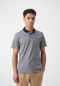 Pilkas polo marškinėlis su juodu apykaklė ir baltais dryžiais. Yra mažas logotipas ant krūtinės. Trumpomis rankovėmis ir klasikinio kirpimo.