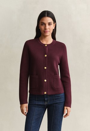 Femme portant un cardigan bordeaux à manches longues, boutonné avec des poches avant et un jean bleu foncé ajusté, debout devant un fond uni.