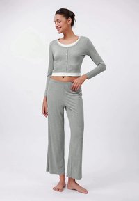 Jauna moteris šypsosi, vilkėdama šviesiai pilką trumpą kardiganą ir atitinkančias plačias kelnes, stovėdama basi prieš neutralų foną.