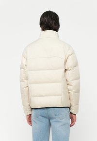 Homme portant une doudoune couleur crème et un jean bleu clair, se tenant de dos contre un fond blanc uni.