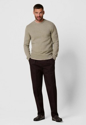 Man draagt een beige trui, zwarte broek en zwarte schoenen, staat met handen in de zakken tegen een effen grijze achtergrond.