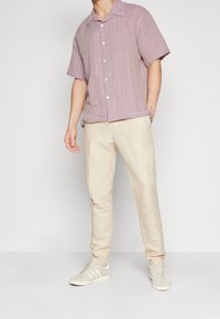 Korte mouwen, overhemd met kraag in lichtpaars met een geruit patroon; gecombineerd met beige slim-fit broek en grijze sneakers met witte strepen.