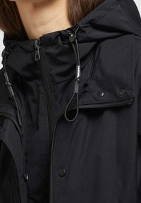 Lähikuva mustasta hupullisesta takista, jossa on säädettävät kiristysnauhat, vetoketjusuljin ja painonapillinen läppä henkilön ylävartalolla.