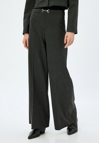 Graue, weit geschnittene Hosen aus strukturiertem Material, mit einer glänzenden silbernen Hardware-Akzent am Bund. Abgerundet mit schwarzen spitzen Schuhen.