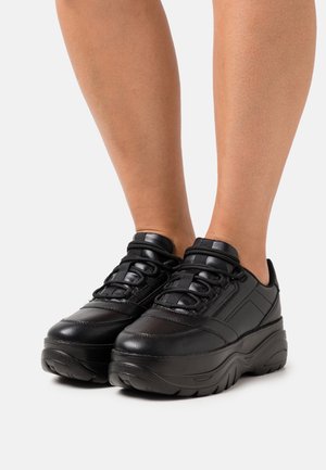 Zapatillas negras con suela gruesa, con parte superior de cuero liso, punta redonda y cordones gruesos. Sutiles acentos acolchados en los laterales.
