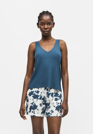 Femme portant un top bleu sans manches à col en V et un short blanc avec un imprimé floral bleu foncé, debout devant un fond clair uni.