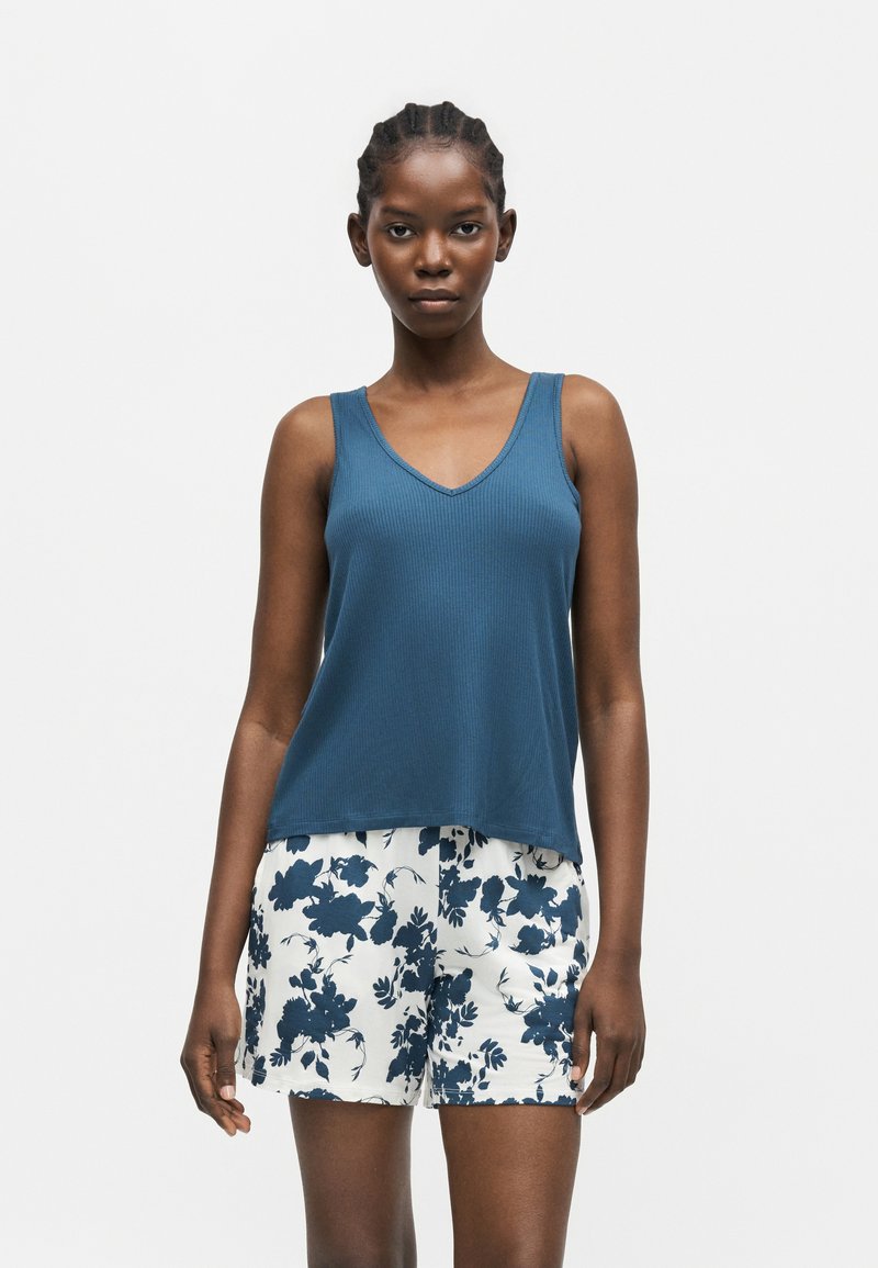 Femme portant un top bleu sans manches à col en V et un short blanc avec un imprimé floral bleu foncé, debout devant un fond clair uni.