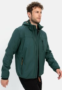 Grøn, vandtæt jakke med lynlås foran, hætte og flere lommer. Har sorte detaljer og en tekstureret overflade.