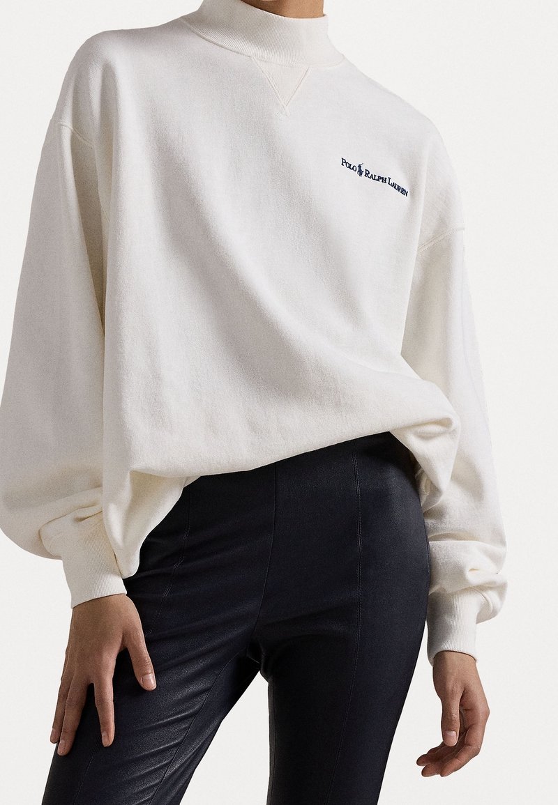 Nainen, joka on pukeutunut tilavaan valkoiseen Polo Ralph Lauren -huppariin ja tiukkoihin mustiin nahkahousuihin, seisoo kädet rentoina sivuilla.