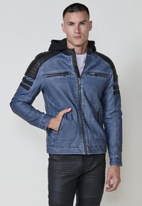 Uomo con giacca blu, con pannelli spallati neri imbottiti e cerniere, che posa con una mano in tasca su uno sfondo semplice.