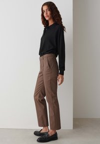 Bruine rechte broek met voorplooien, gecombineerd met een zwarte poloshirt met lange mouwen. De outfit wordt compleet gemaakt met zwarte instapschoenen.