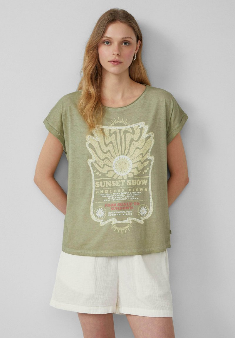 T-shirt à manches courtes vert olive avec un design graphique blanc représentant un soleil et le texte "SUNSET SHOW" sur le devant ; coupe décontractée, texture douce.