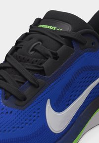 Μπλε αθλητικό παπούτσι Nike με μαύρα κορδόνια, λευκό λογότυπο Swoosh και φωσφορίζουσες πράσινες λεπτομέρειες στη σόλα και στη γλώσσα του παπουτσιού.