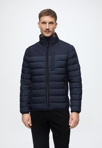 Tengerészkék puffertájú dzseki, steppelt kialakítással, magas gallérral, elülső cipzárral és mellzsebbe. Simás textúra, testhezálló forma és oldalsó cipzáras zsebek.