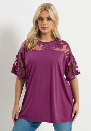 Kvinde iført en lilla blomstret blondebluse med gennemsigtige ærmer og bærestykke, kombineret med blå jeans, stående foran en ensfarvet baggrund.